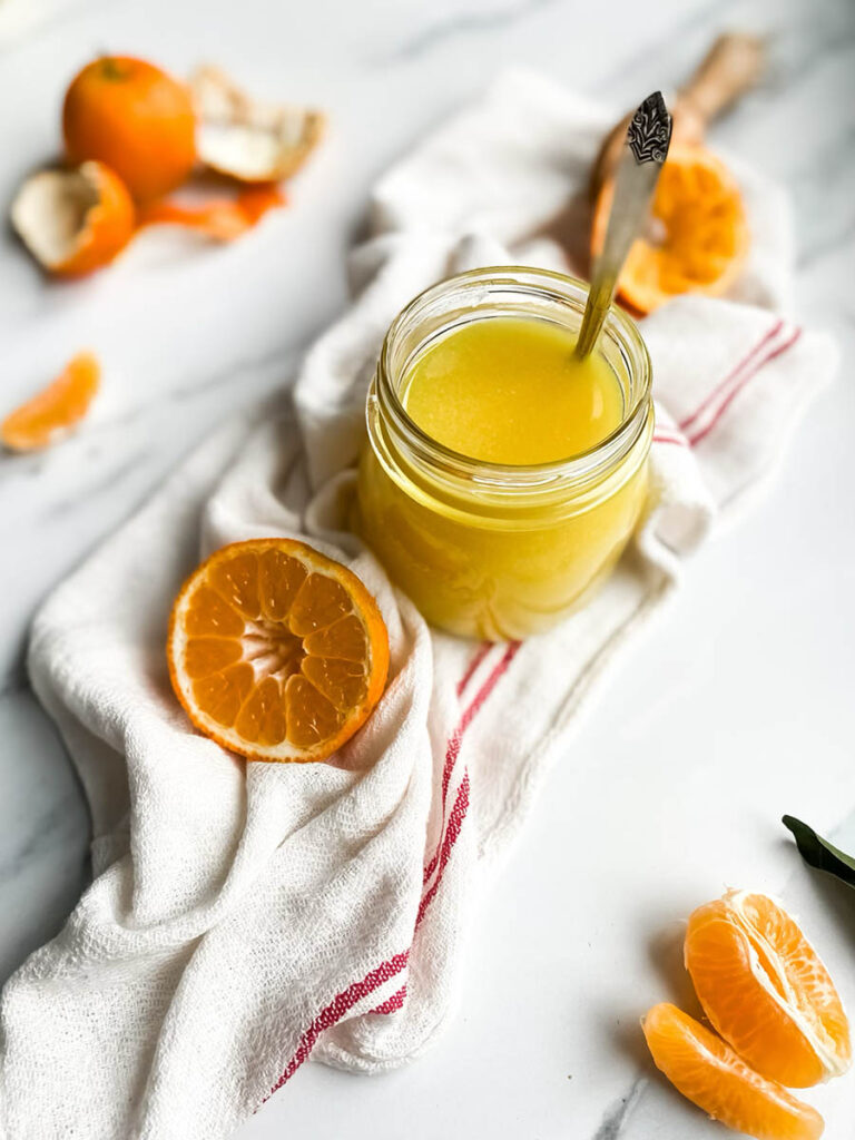 A jar of bright orange clementine curd with a spoon, resting on a white cloth with red stripes. Fresh clementine slices and peeled segments are scattered around, along with a wooden citrus reamer, creating a fresh and vibrant setting. The background is a clean white marble surface, emphasizing the homemade citrus spread.