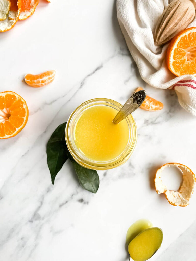 A jar of bright orange clementine curd with a spoon, resting on a white cloth with red stripes. Fresh clementine slices and peeled segments are scattered around, along with a wooden citrus reamer, creating a fresh and vibrant setting. The background is a clean white marble surface, emphasizing the homemade citrus spread.