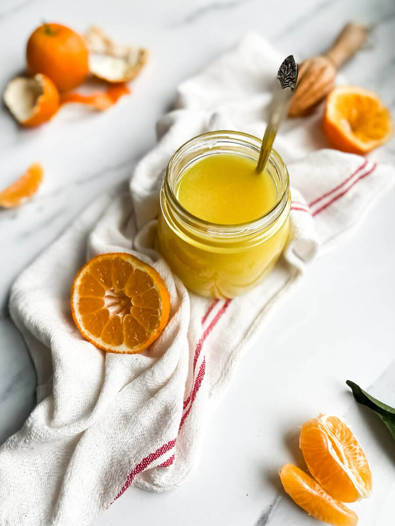 A jar of bright orange clementine curd with a spoon, resting on a white cloth with red stripes. Fresh clementine slices and peeled segments are scattered around, along with a wooden citrus reamer, creating a fresh and vibrant setting. The background is a clean white marble surface, emphasizing the homemade citrus spread.