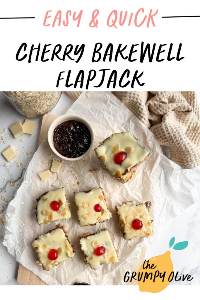 Cherry Bakewell Flapjack, Quick & Delicious Recipe 9 Pin image for the cherry bakewell flapjack. An overhead view of cherry bakewell flapjack arranged on a wooden board lined with parchment paper. The golden-baked squares are topped with white icing, scattered with almonds, and garnished with bright red cherries. A jar of oats, white chocolate chunks, and a bowl of dark jam sit nearby on a marble surface, accompanied by a textured beige cloth.