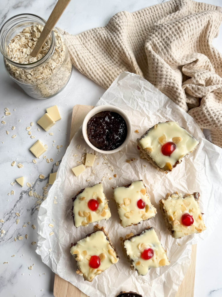 Cherry Bakewell Flapjack, Quick & Delicious Recipe 6 An overhead view of cherry bakewell flapjack arranged on a wooden board lined with parchment paper. The golden-baked squares are topped with white icing, scattered with almonds, and garnished with bright red cherries. A jar of oats, white chocolate chunks, and a bowl of dark jam sit nearby on a marble surface, accompanied by a textured beige cloth.