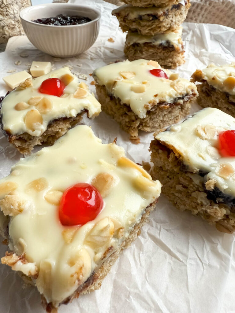Cherry Bakewell Flapjack, Quick & Delicious Recipe 3 A close-up of cherry bakewell flapjack showcasing their crumbly, golden oat base topped with creamy white icing, almond flakes, and red cherries. In the background, a stack of flapjacks and a small bowl of dark jam add depth to the presentation on the parchment-covered surface.