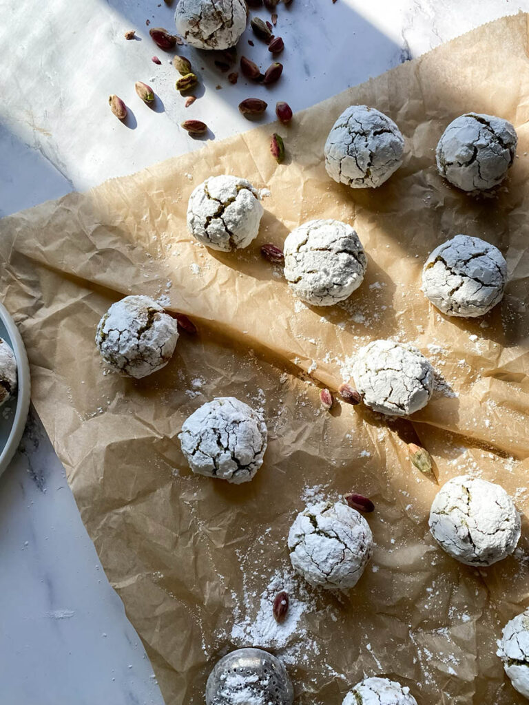 Yummy Pistachio Amaretti Cookies, For The Most Perfect Christmas 1 Multiple pistachio amaretti cookies, dusted with powdered sugar, are arranged on parchment paper with a few whole pistachios scattered around. The image showcases the rustic, crinkled texture of the cookies.