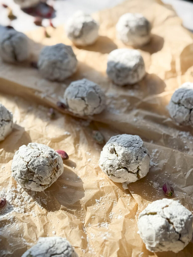 Yummy Pistachio Amaretti Cookies, For The Most Perfect Christmas 8 Multiple pistachio amaretti cookies, dusted with powdered sugar, are arranged on parchment paper with a few whole pistachios scattered around. The image showcases the rustic, crinkled texture of the cookies.