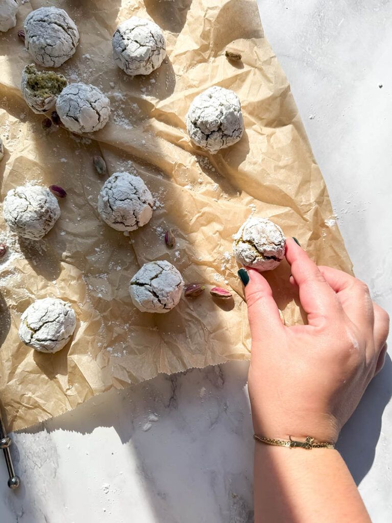 Yummy Pistachio Amaretti Cookies, For The Most Perfect Christmas 9 A hand reaching for a cracked, powdered pistachio amaretti cookie on a sheet of brown parchment paper. The scene is illuminated by natural light, highlighting several cookies and scattered pistachios around.