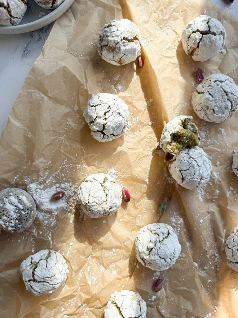 Yummy Pistachio Amaretti Cookies, For The Most Perfect Christmas 2 Multiple pistachio amaretti cookies, dusted with powdered sugar, are arranged on parchment paper with a few whole pistachios scattered around. The image showcases the rustic, crinkled texture of the cookies.