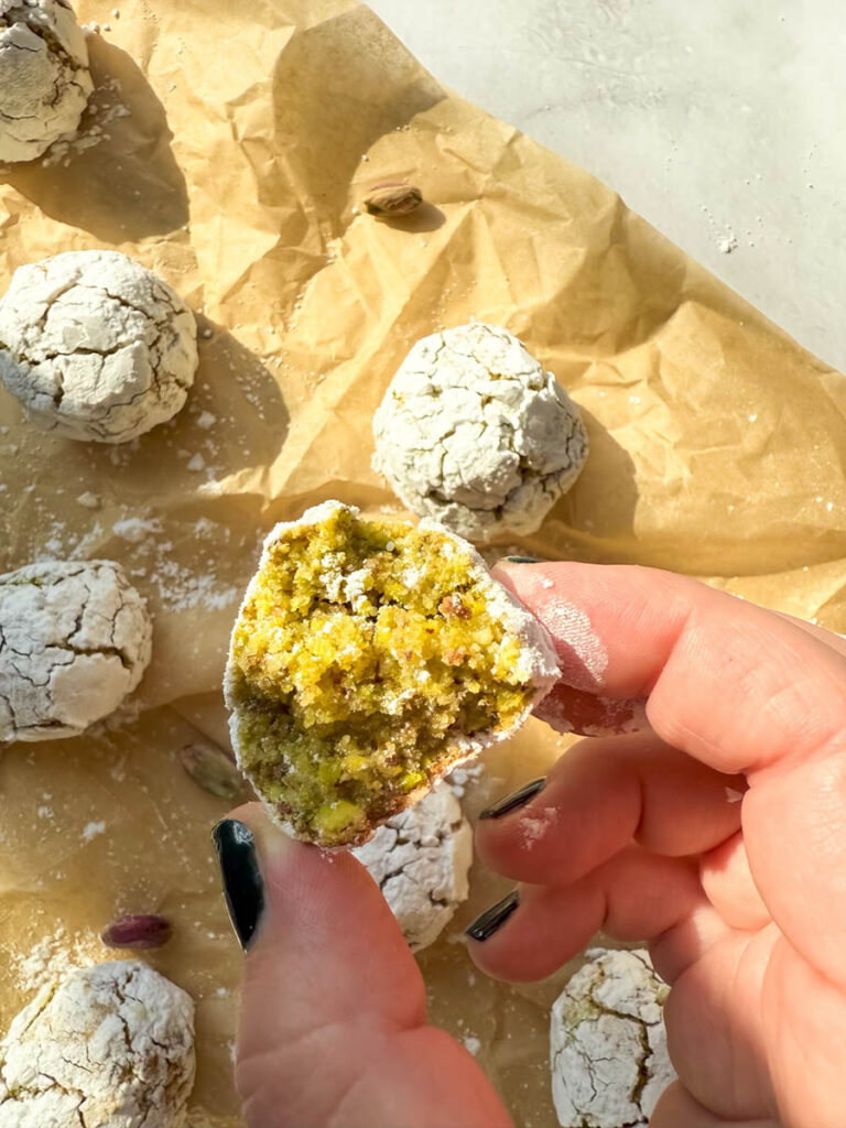 Yummy Pistachio Amaretti Cookies, For The Most Perfect Christmas 3 A close-up of a hand holding a half-eaten pistachio amaretti cookie, showing its vibrant green interior. The cookie is covered in a thin layer of powdered sugar, and additional cookies rest on parchment paper in the background.