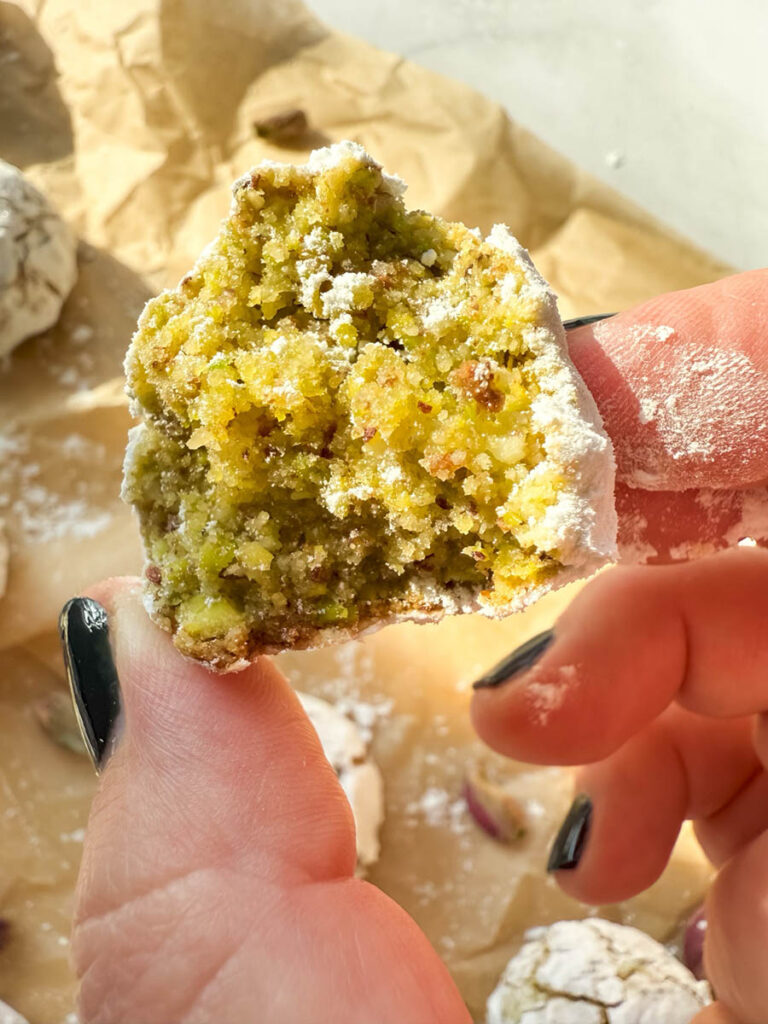 Yummy Pistachio Amaretti Cookies, For The Most Perfect Christmas 4 A close-up of a hand holding a half-eaten pistachio amaretti cookie, showing its vibrant green interior. The cookie is covered in a thin layer of powdered sugar, and additional cookies rest on parchment paper in the background.