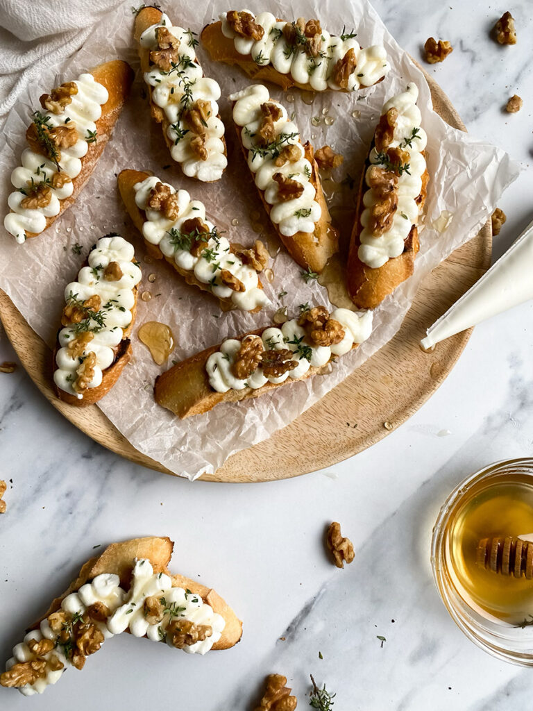A wooden platter topped with toasted ricotta honey crostini drizzled with honey, piped ricotta, walnuts, and a sprinkle of fresh thyme, resting on parchment paper. A honey dipper and scattered walnuts lie nearby on a marble surface.