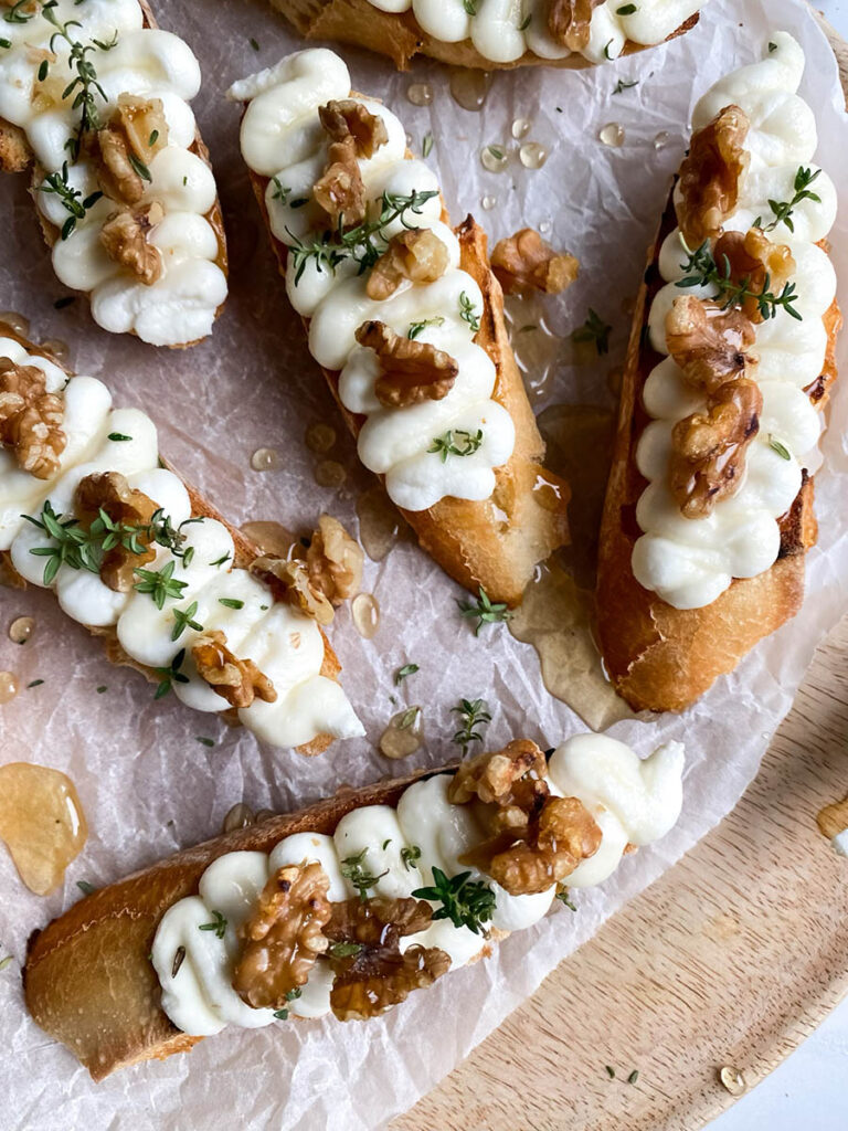 A wooden platter topped with toasted ricotta honey crostini drizzled with honey, piped ricotta, walnuts, and a sprinkle of fresh thyme, resting on parchment paper. A honey dipper and scattered walnuts lie nearby on a marble surface.