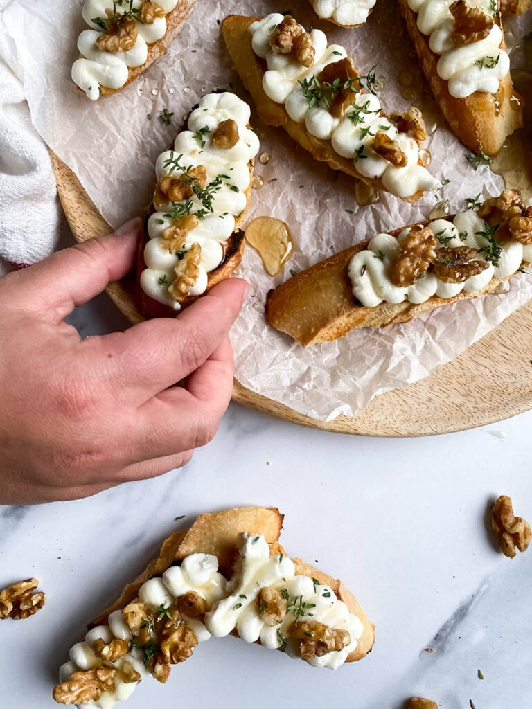 A hand reaching for a ricotta honey crostini from a wooden platter. The crostini is decorated with honey, walnuts, and thyme, with a white cloth adding texture to the scene.