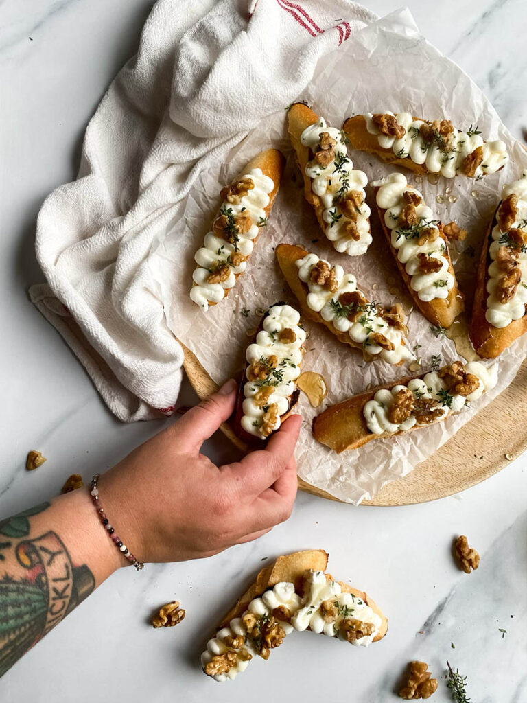 A hand reaching for a ricotta honey crostini from a wooden platter. The crostini is decorated with honey, walnuts, and thyme, with a white cloth adding texture to the scene.