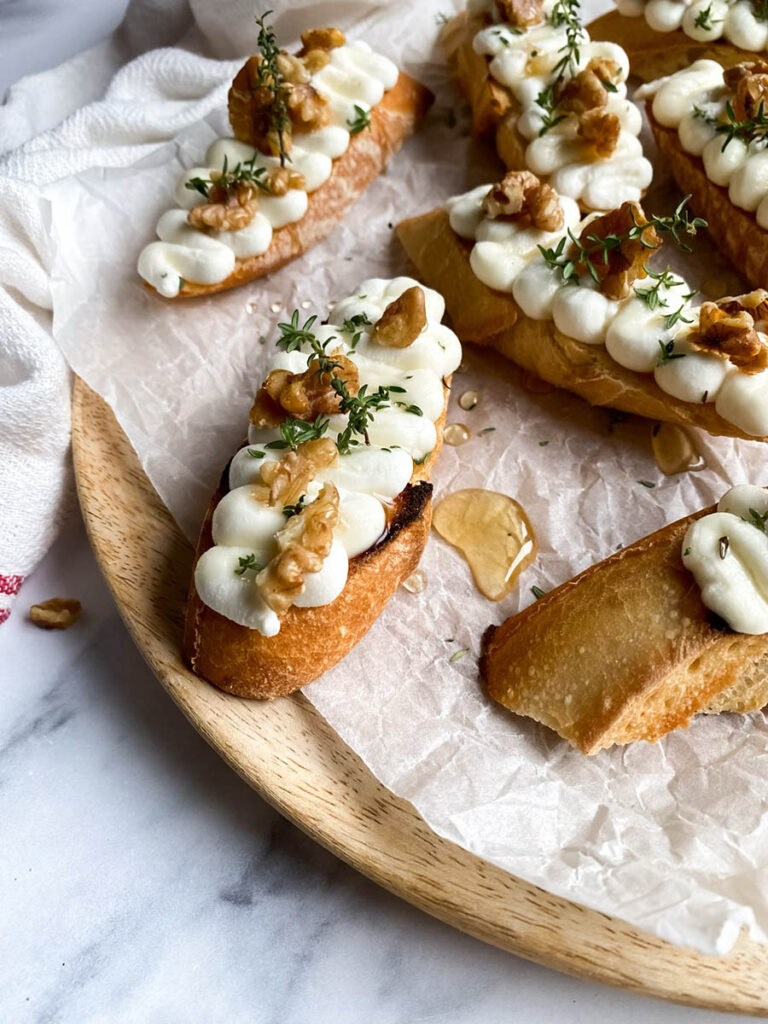 A wooden platter topped with toasted ricotta honey crostini drizzled with honey, piped ricotta, walnuts, and a sprinkle of fresh thyme, resting on parchment paper. A honey dipper and scattered walnuts lie nearby on a marble surface.