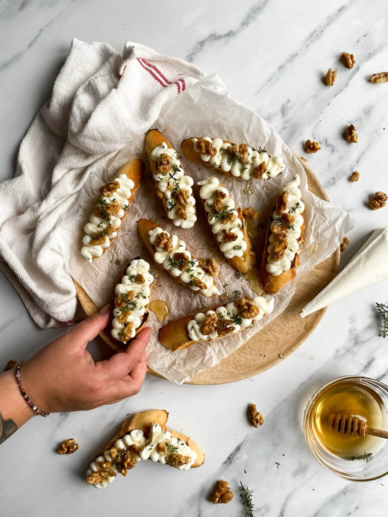 A hand reaching for a ricotta honey crostini from a wooden platter. The crostini is decorated with honey, walnuts, and thyme, with a white cloth adding texture to the scene.