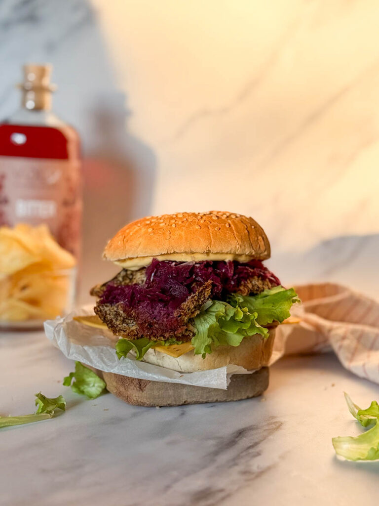 Asian-Inspired Oyster Mushroom Burgers, Easy & Vegan Recipe 2 A golden, crispy vegan oyster mushroom burger stacked with fresh lettuce, cheese, and purple coleslaw, resting on parchment paper on a marble surface, surrounded by soft, natural light and a blurred background of condiments and chips.