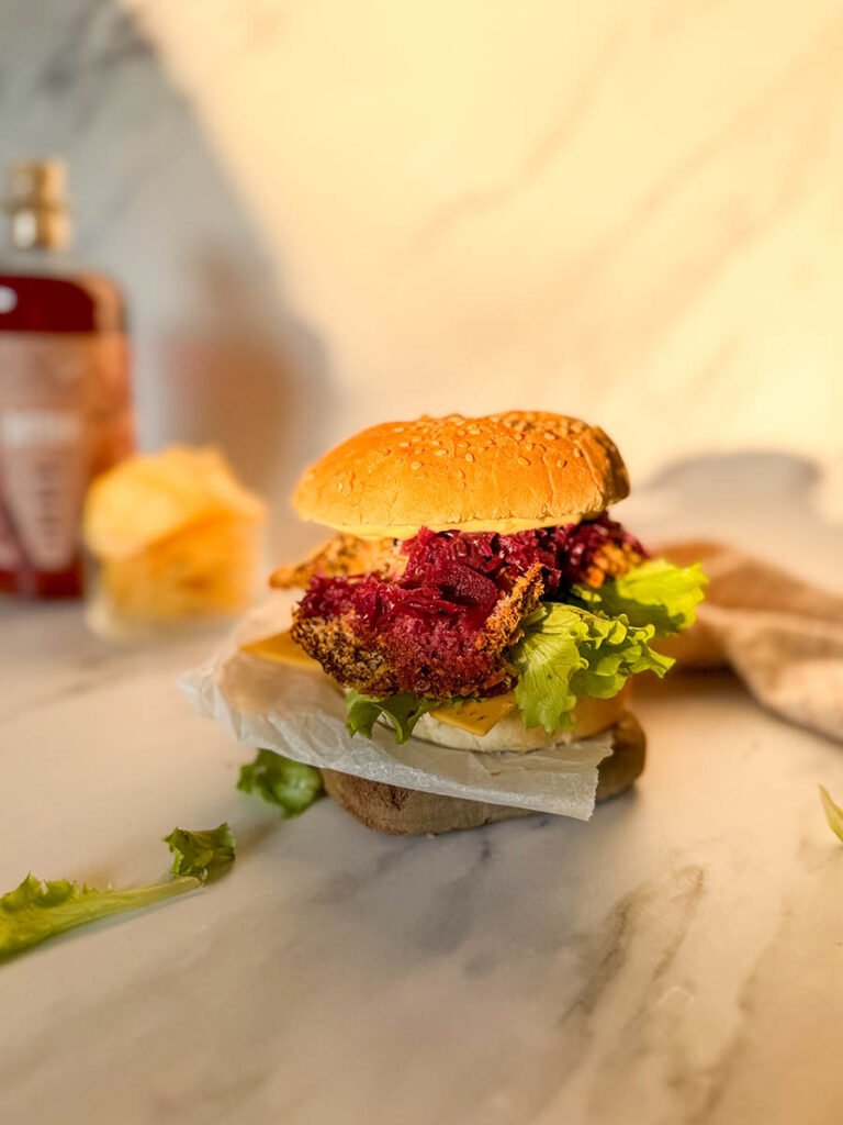 Asian-Inspired Oyster Mushroom Burgers, Easy & Vegan Recipe 3 A golden, crispy vegan oyster mushroom burger stacked with fresh lettuce, cheese, and purple coleslaw, resting on parchment paper on a marble surface, surrounded by soft, natural light and a blurred background of condiments and chips.