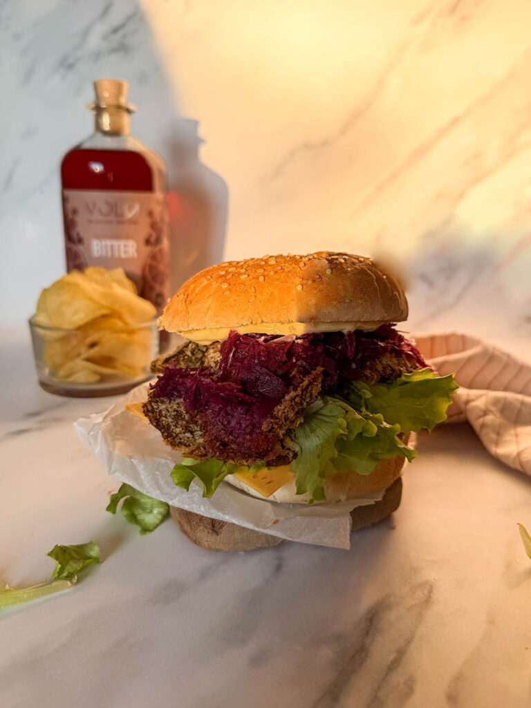 Asian-Inspired Oyster Mushroom Burgers, Easy & Vegan Recipe 6 A close-up view of vegan oyster mushroom burgers featuring a crispy breaded mushroom patty, vibrant purple coleslaw, fresh lettuce, and cheese layered on a sesame seed bun, with a blurred background including chips and a sauce bottle.