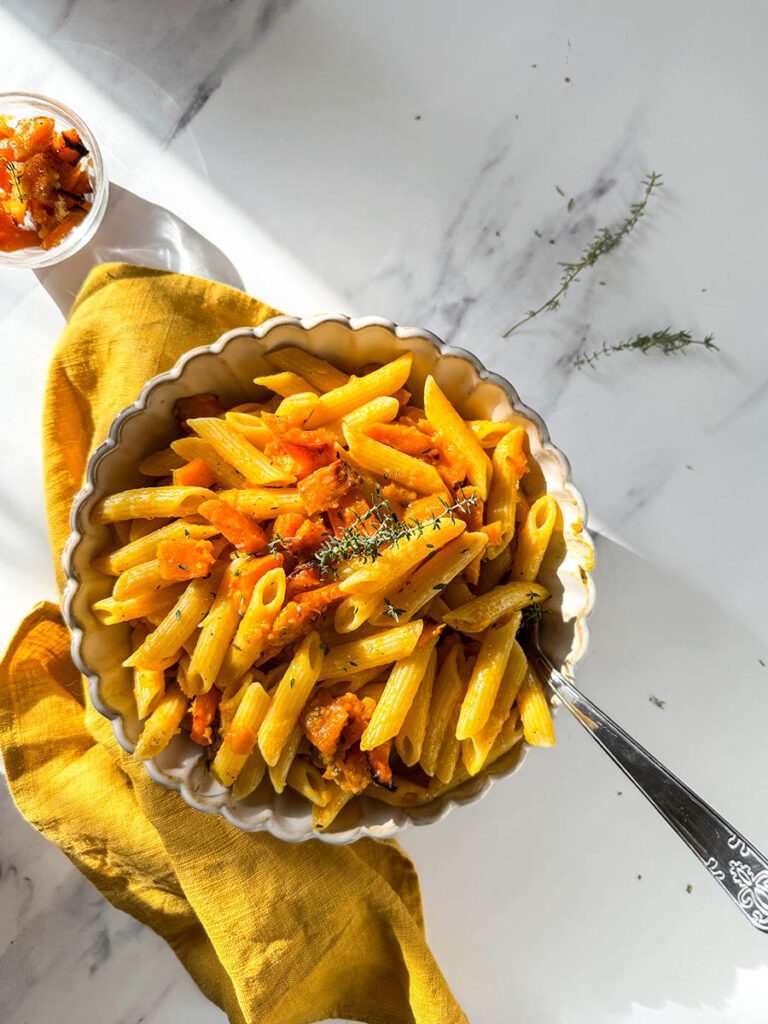 A close-up of vegan pumpkin feta pasta, featuring penne coated in a creamy pumpkin sauce with roasted pumpkin pieces and fresh thyme, served in a scalloped bowl on a yellow cloth. Sunlight enhances the vibrant yellow tones on a white marble surface.