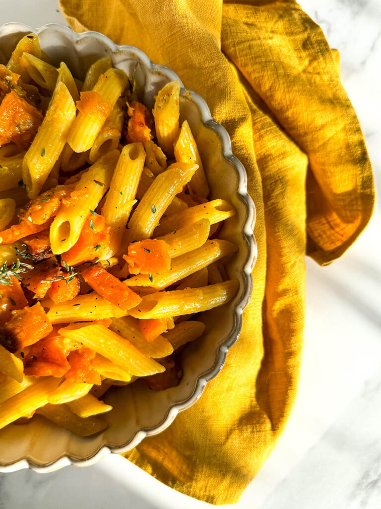 A bowl of vegan pumpkin feta pasta with penne, roasted pumpkin chunks, and a hint of fresh herbs, placed on a mustard yellow cloth. The dish’s warm tones are highlighted by natural light against the white marble background.