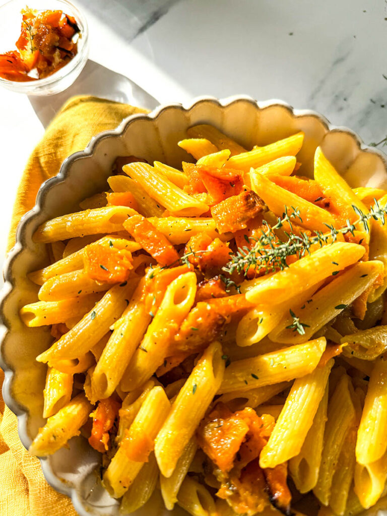 Vegan pumpkin feta pasta in a decorative bowl, showcasing golden penne pasta with roasted pumpkin and thyme. The bright sunlight highlights the textures and colors of the pasta on a white marble table.