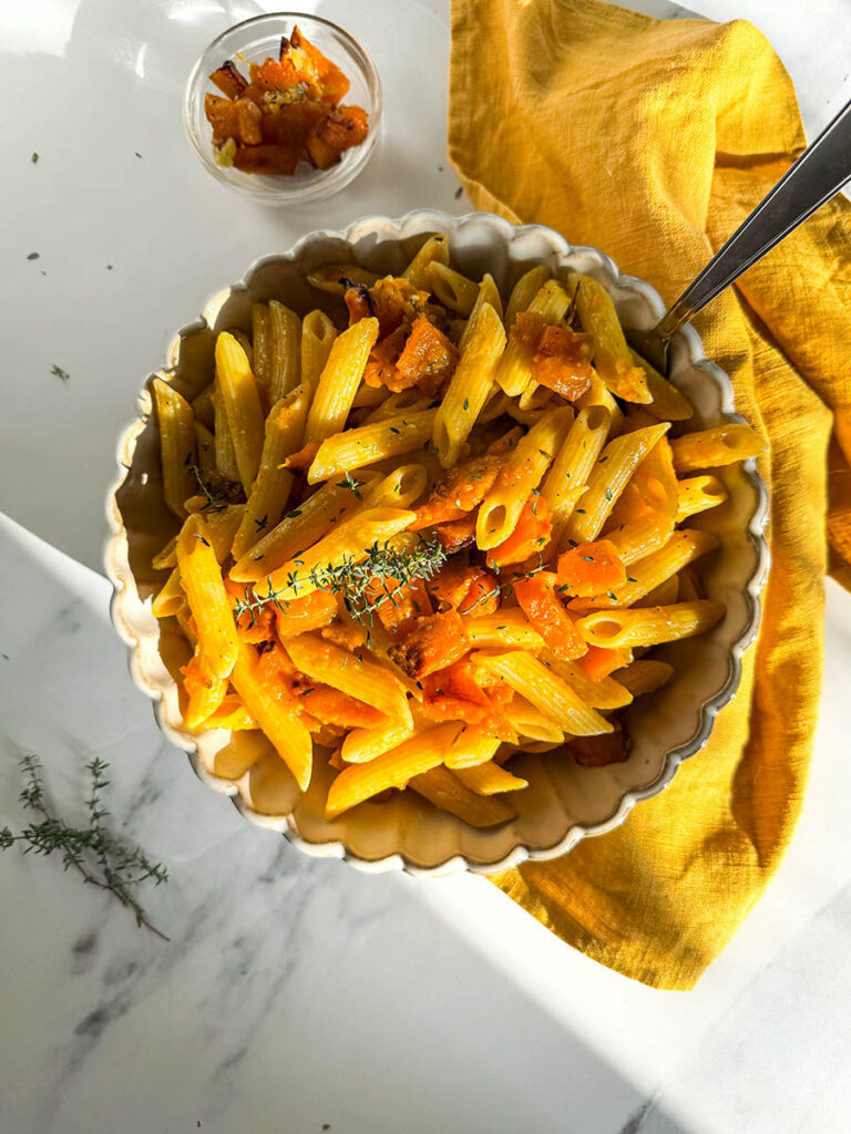 A bowl of vegan pumpkin feta pasta with penne, roasted pumpkin chunks, and a hint of fresh herbs, placed on a mustard yellow cloth. The dish’s warm tones are highlighted by natural light against the white marble background.