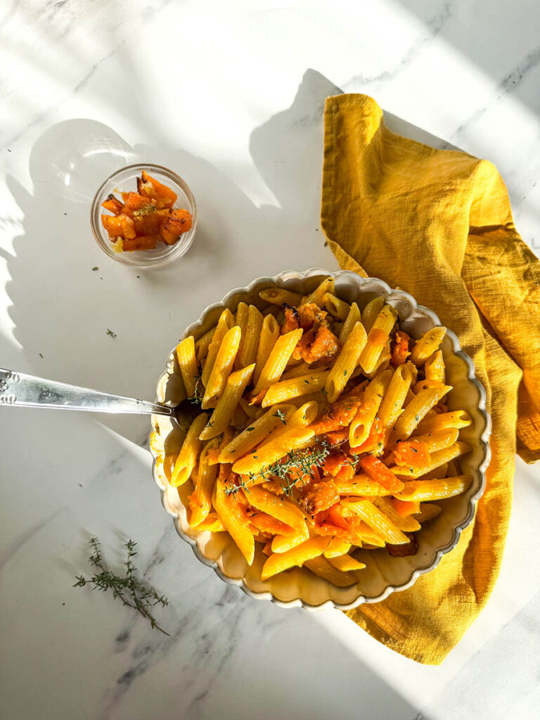 A bowl of vegan pumpkin feta pasta with penne, roasted pumpkin chunks, and a hint of fresh herbs, placed on a mustard yellow cloth. The dish’s warm tones are highlighted by natural light against the white marble background.
