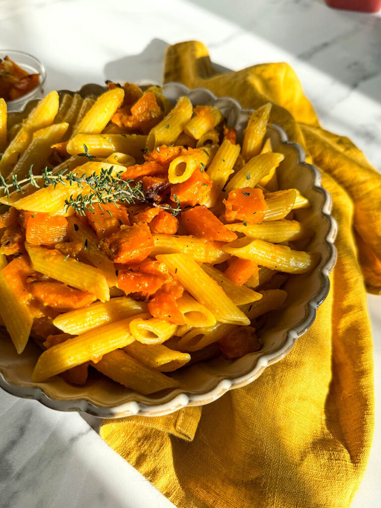 A bowl of vegan pumpkin feta pasta with penne, roasted pumpkin chunks, and a hint of fresh herbs, placed on a mustard yellow cloth. The dish’s warm tones are highlighted by natural light against the white marble background.