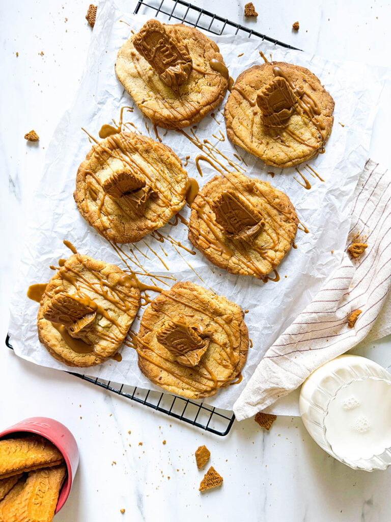 Vegan Biscoff Cookies, The Ultimate Recipe For Delicious Cookies 1 A batch of vegan Biscoff cookies on a black cooling rack, drizzled with caramel-like Biscoff spread and topped with small Biscoff biscuit pieces, placed on a sheet of white parchment paper. Crumbs are scattered on the marble surface, with a cup of milk and a pink mug filled with extra Biscoff biscuits nearby.