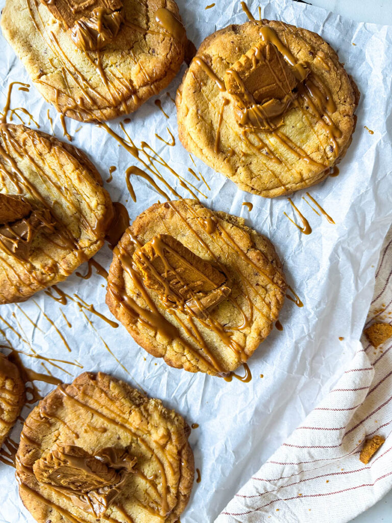 Vegan Biscoff Cookies, The Ultimate Recipe For Delicious Cookies 3 Close-up of several vegan Biscoff cookies drizzled with golden Biscoff spread and topped with pieces of Biscoff biscuits, arranged on parchment paper. The textured cookies show a golden-brown bake, with crumbs and caramel drizzle highlighting the detail.