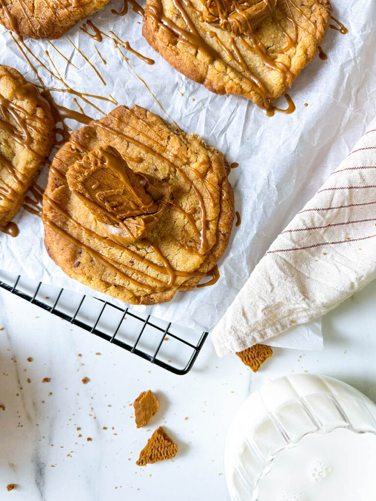 Vegan Biscoff Cookies, The Ultimate Recipe For Delicious Cookies 7 A single vegan Biscoff cookie in focus on a parchment-lined cooling rack, topped with a Biscoff biscuit piece and caramel drizzle. Surrounding the cookie are crumbs and a nearby glass of milk, with a striped cloth napkin adding texture to the minimalist setting.