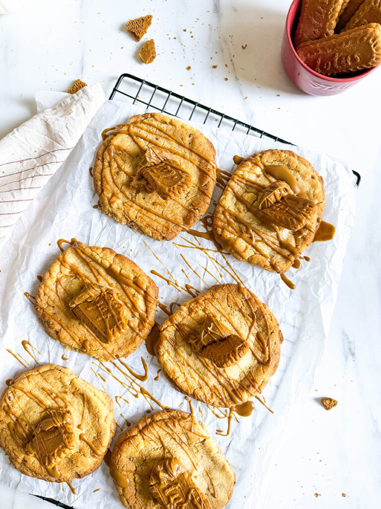 Vegan Biscoff Cookies, The Ultimate Recipe For Delicious Cookies 4 A batch of vegan Biscoff cookies on a black cooling rack, drizzled with caramel-like Biscoff spread and topped with small Biscoff biscuit pieces, placed on a sheet of white parchment paper. Crumbs are scattered on the marble surface, with a cup of milk and a pink mug filled with extra Biscoff biscuits nearby.
