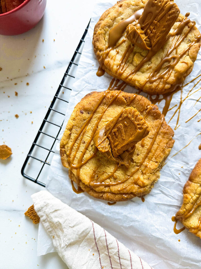 Vegan Biscoff Cookies, The Ultimate Recipe For Delicious Cookies 9 A single vegan Biscoff cookie in focus on a parchment-lined cooling rack, topped with a Biscoff biscuit piece and caramel drizzle. Surrounding the cookie are crumbs and a nearby glass of milk, with a striped cloth napkin adding texture to the minimalist setting.
