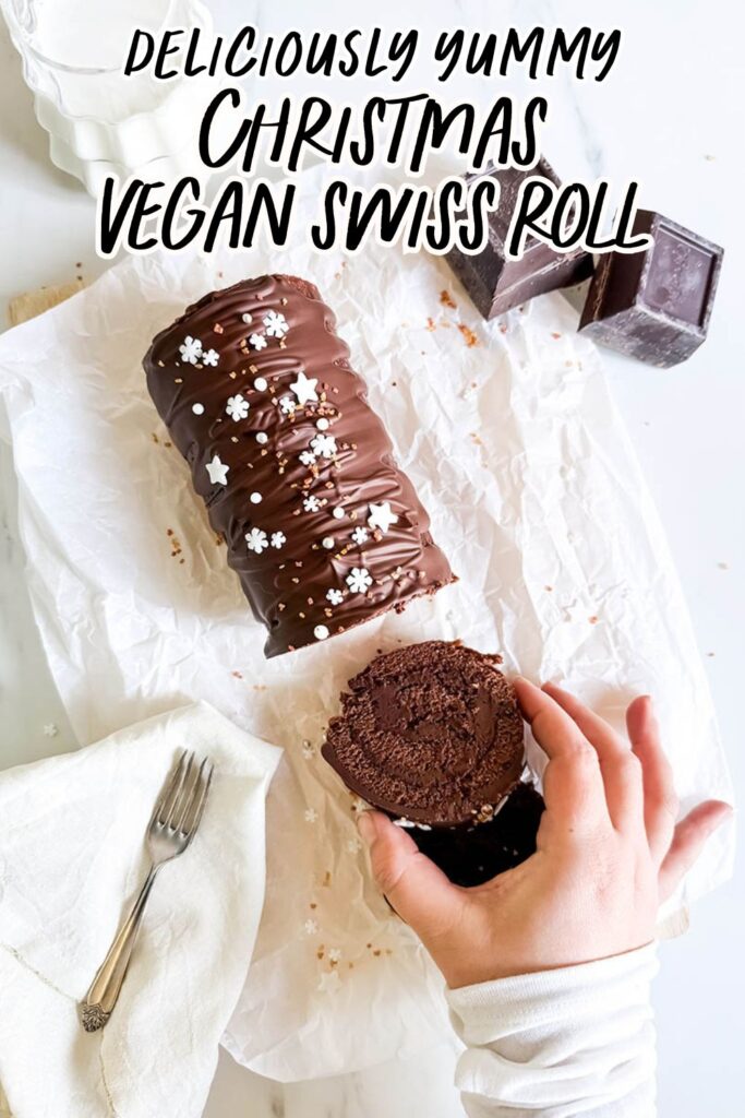 Christmas Vegan Swiss Roll, Easy & Delicious 10 A chocolate Swiss roll covered in glossy chocolate ganache and decorated with small white star and snowflake sprinkles sits on parchment paper. A hand is holding a sliced piece of the roll, revealing its rich, moist texture. In the background are two blocks of dark chocolate and a milk pitcher.