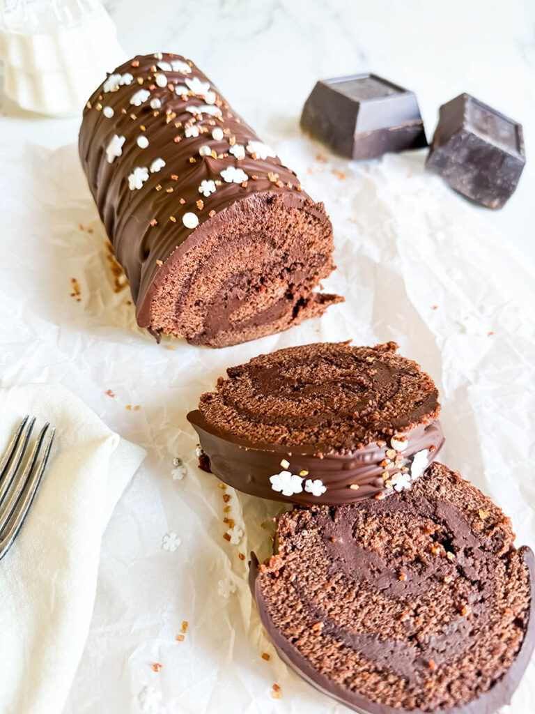 Christmas Vegan Swiss Roll, Easy & Delicious 2 A close-up of a chocolate Swiss roll sliced to reveal its soft, chocolatey swirl interior. The roll is topped with chocolate ganache and festive white and gold sprinkles. A silver fork and cream-colored napkin rest nearby.