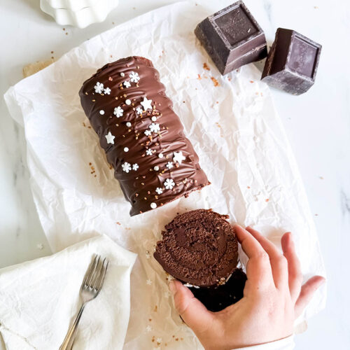 from top- hand reaching for a slice of christmas swiss roll on parchment paper, chocolate and a fork on the sides, milk glass behind, on a marble background