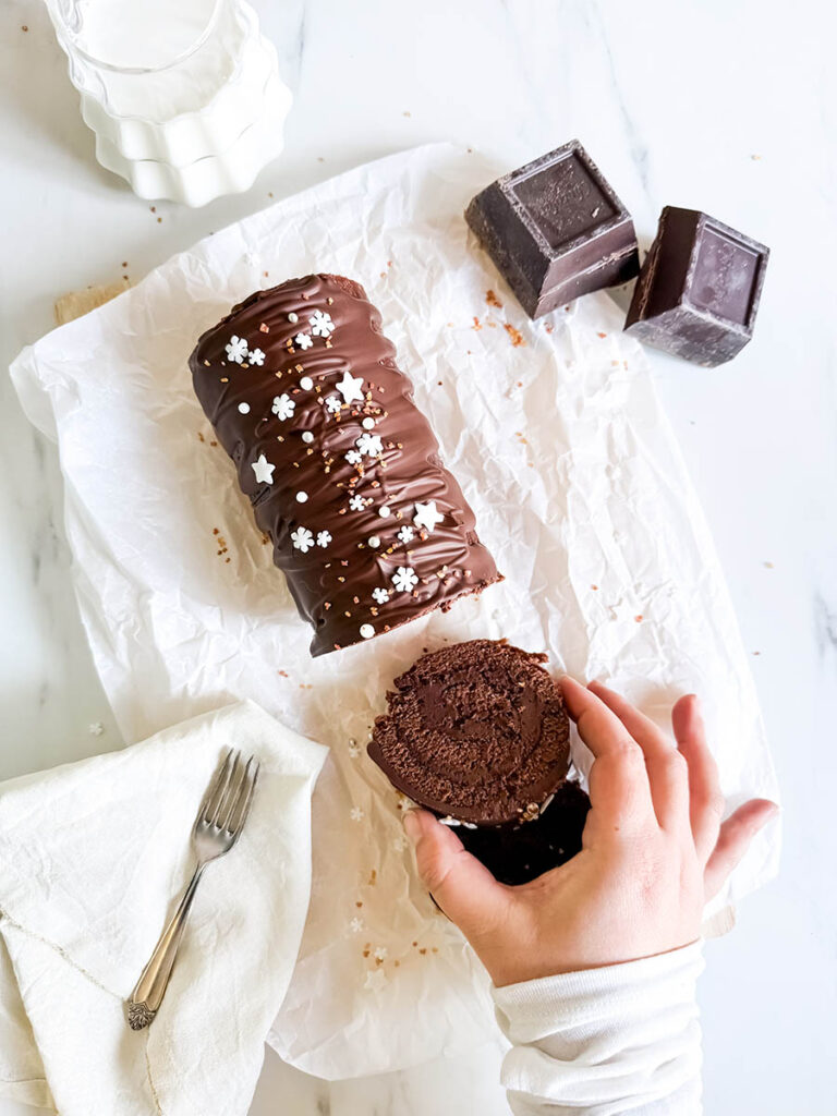 Christmas Vegan Swiss Roll, Easy & Delicious 4 A chocolate Swiss roll covered in glossy chocolate ganache and decorated with small white star and snowflake sprinkles sits on parchment paper. A hand is holding a sliced piece of the roll, revealing its rich, moist texture. In the background are two blocks of dark chocolate and a milk pitcher.