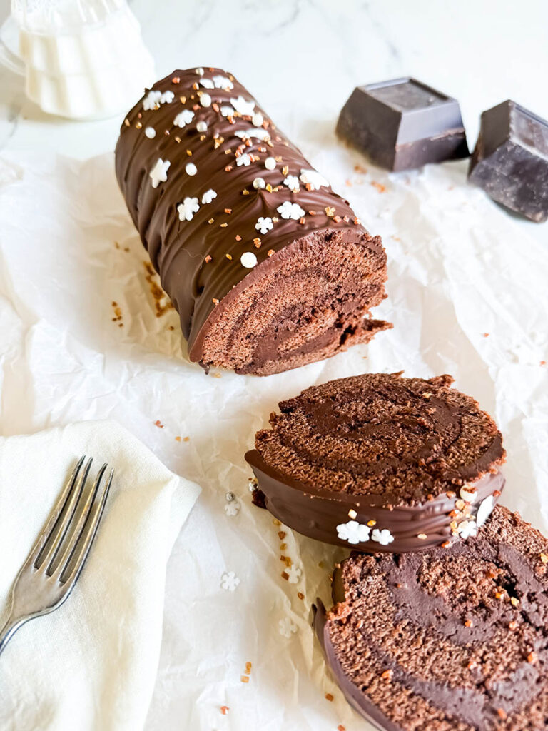 Christmas Vegan Swiss Roll, Easy & Delicious 3 A close-up of a chocolate Swiss roll sliced to reveal its soft, chocolatey swirl interior. The roll is topped with chocolate ganache and festive white and gold sprinkles. A silver fork and cream-colored napkin rest nearby.
