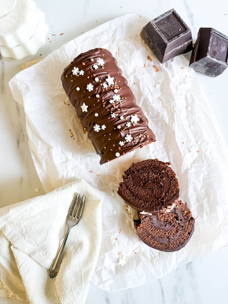 Christmas Vegan Swiss Roll, Easy & Delicious 1 A chocolate Swiss roll displayed on crinkled parchment paper, garnished with white snowflake and star sprinkles. Two slices of the roll lie in front, showcasing the moist interior. A milk pitcher and dark chocolate blocks add to the background ambiance.