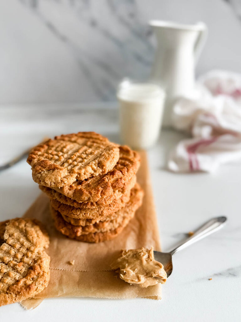 4 Ingredient Peanut Butter Cookies, Easy & Delicious Recipe 2 A close-up of a stack of homemade 4 ingredient peanut butter cookies, with a broken piece in the foreground showing its crumbly texture. A spoonful of peanut butter rests on the parchment paper, and a glass of milk along with a white pitcher adds a cozy background element.