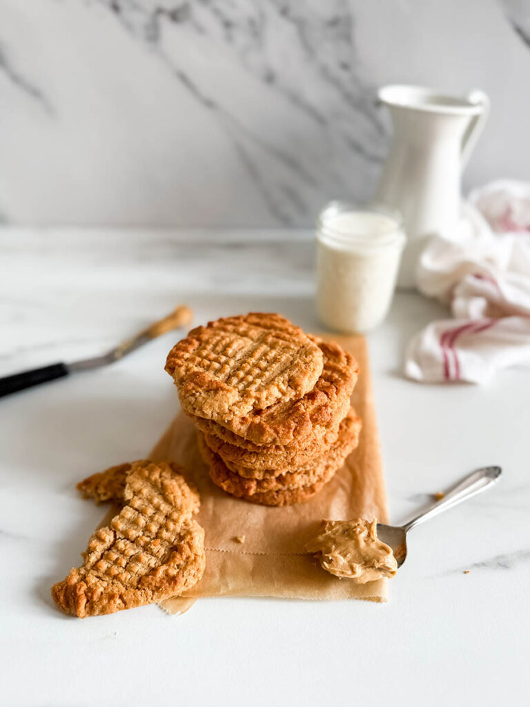 4 Ingredient Peanut Butter Cookies, Easy & Delicious Recipe 1 A close-up of a stack of homemade 4 ingredient peanut butter cookies, with a broken piece in the foreground showing its crumbly texture. A spoonful of peanut butter rests on the parchment paper, and a glass of milk along with a white pitcher adds a cozy background element.