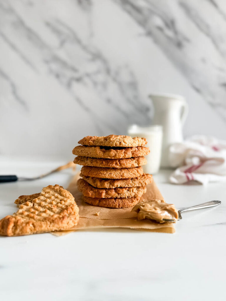4 Ingredient Peanut Butter Cookies, Easy & Delicious Recipe 8 A stack of golden-brown peanut butter cookies sits on a piece of parchment paper, with a broken cookie and a spoonful of peanut butter in the foreground. A glass of milk and a white ceramic pitcher are slightly blurred in the background against a marble-patterned wall.