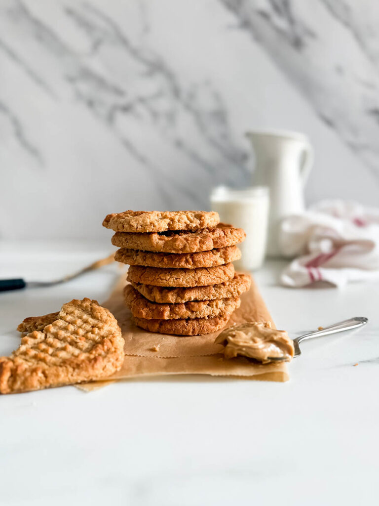 4 Ingredient Peanut Butter Cookies, Easy & Delicious Recipe 10 A stack of golden-brown peanut butter cookies sits on a piece of parchment paper, with a broken cookie and a spoonful of peanut butter in the foreground. A glass of milk and a white ceramic pitcher are slightly blurred in the background against a marble-patterned wall.