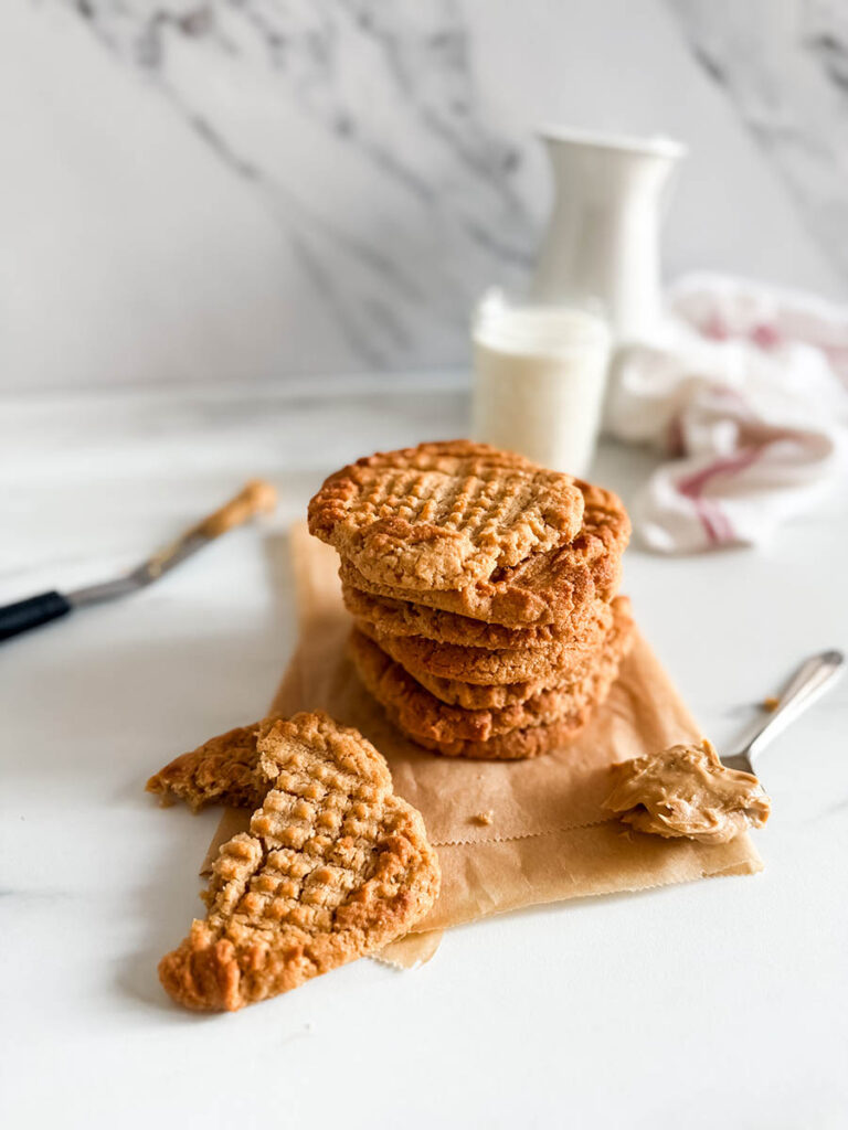 4 Ingredient Peanut Butter Cookies, Easy & Delicious Recipe 4 A stack of golden-brown 4 ingredient peanut butter cookies sits on a piece of parchment paper, with a broken cookie and a spoonful of peanut butter in the foreground. A glass of milk and a white ceramic pitcher are slightly blurred in the background against a marble-patterned wall.