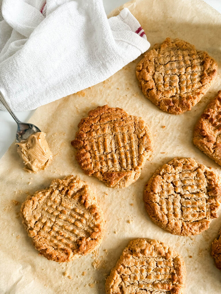 4 Ingredient Peanut Butter Cookies, Easy & Delicious Recipe 9 Freshly baked peanut butter cookies with a classic crisscross pattern sit on parchment paper. A spoonful of creamy peanut butter rests nearby, with a soft, white kitchen towel partially visible in the background.