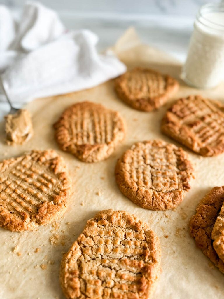 4 Ingredient Peanut Butter Cookies, Easy & Delicious Recipe 5 Freshly baked 4 ingredient peanut butter cookies with a classic crisscross pattern sit on parchment paper. A spoonful of creamy peanut butter rests nearby, with a soft, white kitchen towel partially visible in the background.