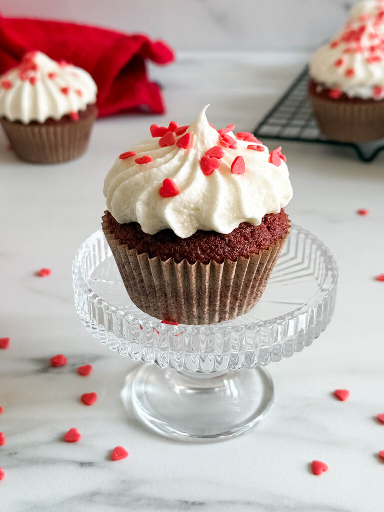 Baileys Red Velvet Cupcakes, The Perfect Valentine's Dessert 1 A single Baileys red velvet cupcake topped with creamy white frosting and heart-shaped red sprinkles, displayed on a clear glass stand, with blurred cupcakes and a red napkin in the background.