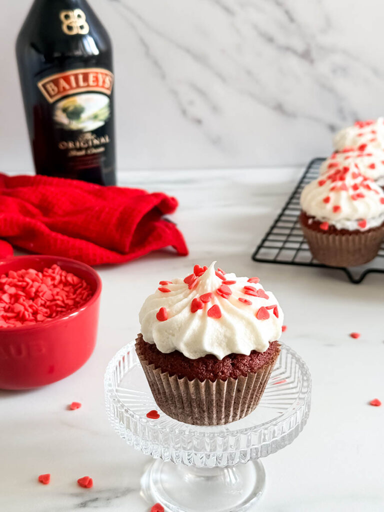 Baileys Red Velvet Cupcakes, The Perfect Valentine's Dessert 5 A close-up of a red velvet cupcake with white frosting and red heart sprinkles, accompanied by a Baileys bottle, a red bowl of sprinkles, and a blurred cupcake cooling rack in the background.