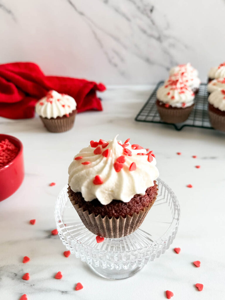 Baileys Red Velvet Cupcakes, The Perfect Valentine's Dessert 2 A Baileys red velvet cupcake decorated with white frosting and heart-shaped red sprinkles, sitting on a glass stand, with additional cupcakes on a cooling rack in the background.