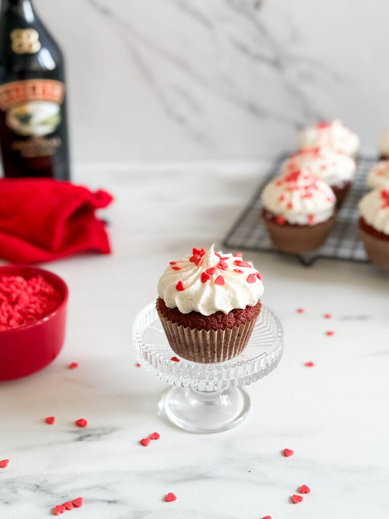 Baileys Red Velvet Cupcakes, The Perfect Valentine's Dessert 3 A Baileys red velvet cupcake with creamy frosting and red heart sprinkles on a glass stand, surrounded by scattered sprinkles, a Baileys bottle, and other cupcakes in the background.