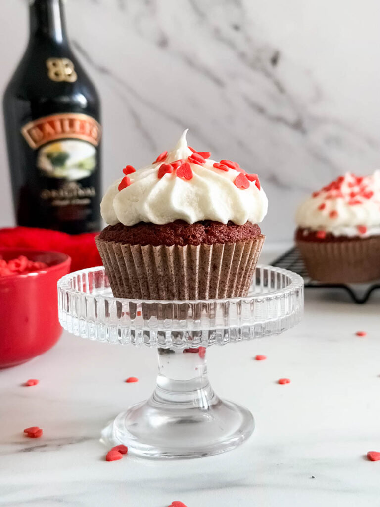 Baileys Red Velvet Cupcakes, The Perfect Valentine's Dessert 4 A Baileys red velvet cupcake with creamy frosting and red heart sprinkles on a glass stand, surrounded by scattered sprinkles, a Baileys bottle, and other cupcakes in the background.